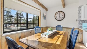 Ein Esszimmer mit Holztisch, blauen Stühlen, einer Uhr an der Wand und einem Fenster mit Blick auf Bäume und parkende Autos.