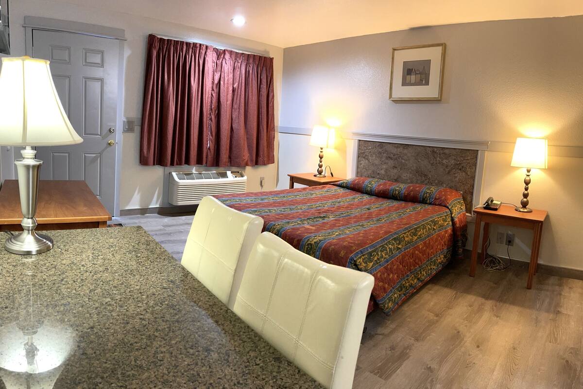 A hotel room with a bed, two bedside lamps, a desk with a lamp, a chair, a window with curtains, and a picture on the wall.