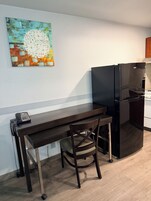 Una cocina compacta con una mesa de madera oscura, una silla, un refrigerador negro y un cuadro colgado en la pared.
