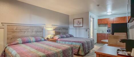 A hotel room with two beds, a wooden headboard, a nightstand with a lamp, a small table, and a kitchenette with a microwave.