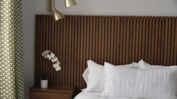 A neatly made bed with white linens, a wooden slatted headboard, a bedside table with a potted plant, and a wall-mounted lamp. - Hall Building (Aledo)