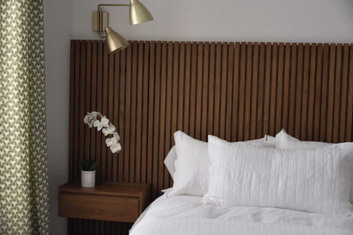 A neatly made bed with white linens, a wooden slatted headboard, a bedside table with a potted plant, and a wall-mounted lamp.