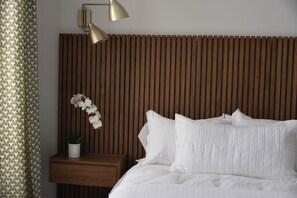 A neatly made bed with white linens, a wooden slatted headboard, a bedside table with a potted plant, and a wall-mounted lamp.
