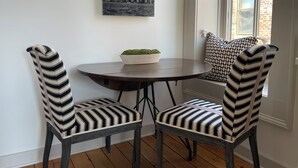 A corner seating area with a round table, two striped chairs, a bowl with green plants, a black and white cityscape painting, and a window with a brick wall. - Hall Building (Aledo)
