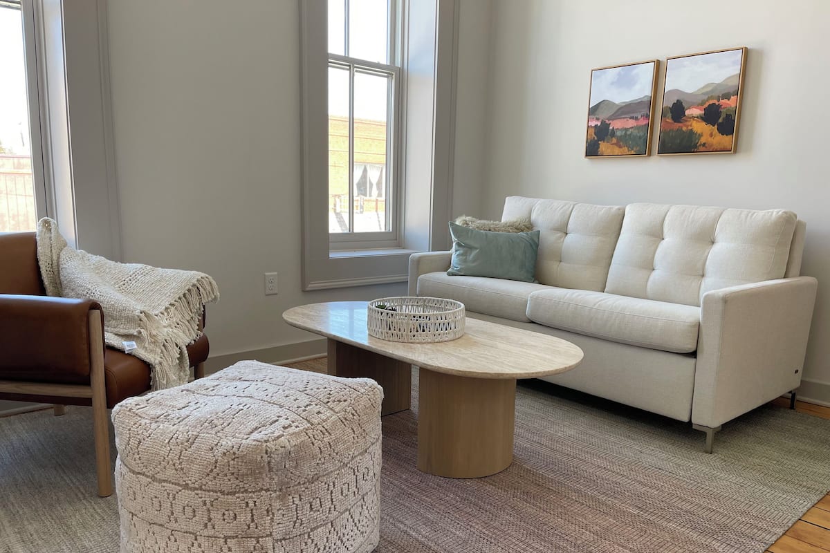 A living room with a brown leather chair, a white sofa, a round coffee table, and a patterned ottoman. There are two windows with curtains, a rug, and two landscape paintings on the wall.