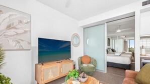 Una sala moderna con un televisor de pantalla plana grande, un mueble de madera, una mesa de centro redonda con objetos decorativos, un sillón verde y una cama en la habitación contigua.