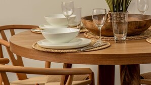 Una mesa de comedor con platos, tazones y vasos blancos, junto con un mantel tejido y una planta en maceta.