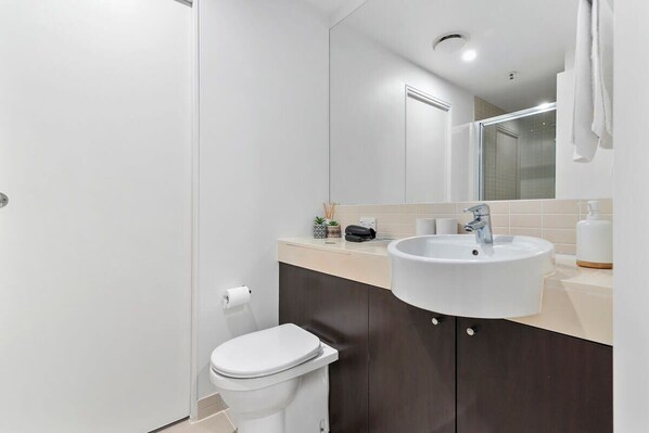 Une salle de bain moderne avec des toilettes blanches, un lavabo surmonté d’un miroir et un meuble de couleur foncée.