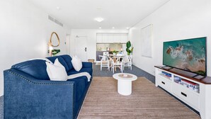 A modern living room with a blue sofa, a coffee table, a TV on a white stand, and a kitchen in the background. - Waterfront Oasis with Pool, Gym and Sauna in Cairns (Cairns City)