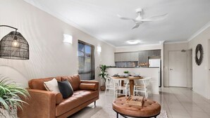 A modern living room with a brown leather sofa, a round ottoman, a dining table with chairs, a kitchen area, and a ceiling fan. - Tropical Retreat with 9 Pools Gym and BBQ Access (Westcourt)