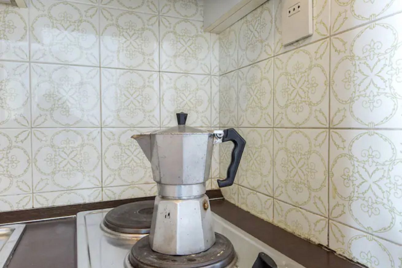A kitchen with a coffee maker on a stove, tiled walls with a floral pattern, and a power outlet above the counter.