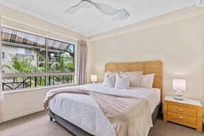 A bedroom with a large window, a bed with a headboard, a bedside table, and a nightstand. - Tropical Resort Apartment with 9 Pools and Gym (Westcourt)