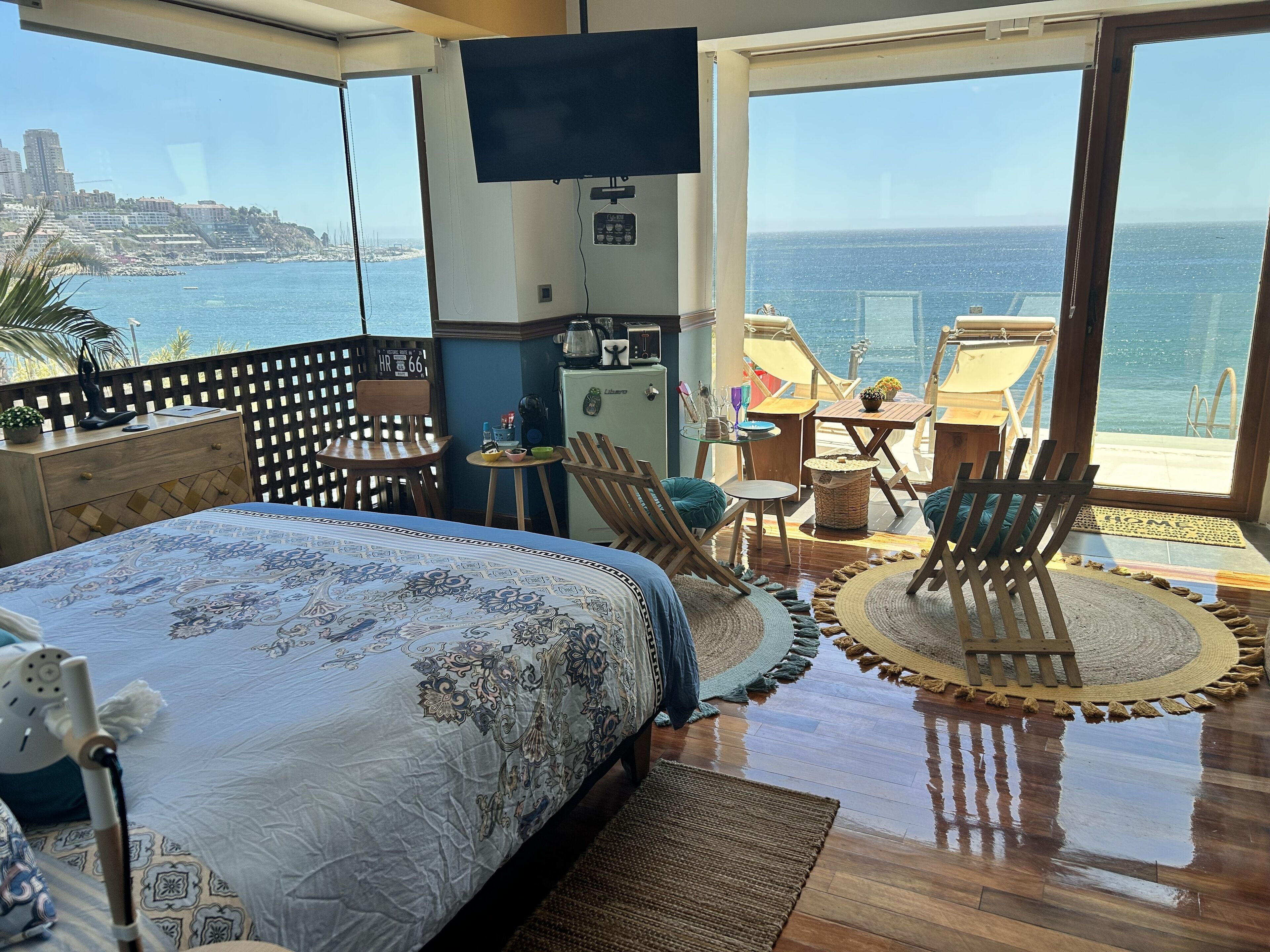 Un dormitorio con una cama grande, un televisor, una mesita y sillas. Ofrece vistas al mar y a edificios.