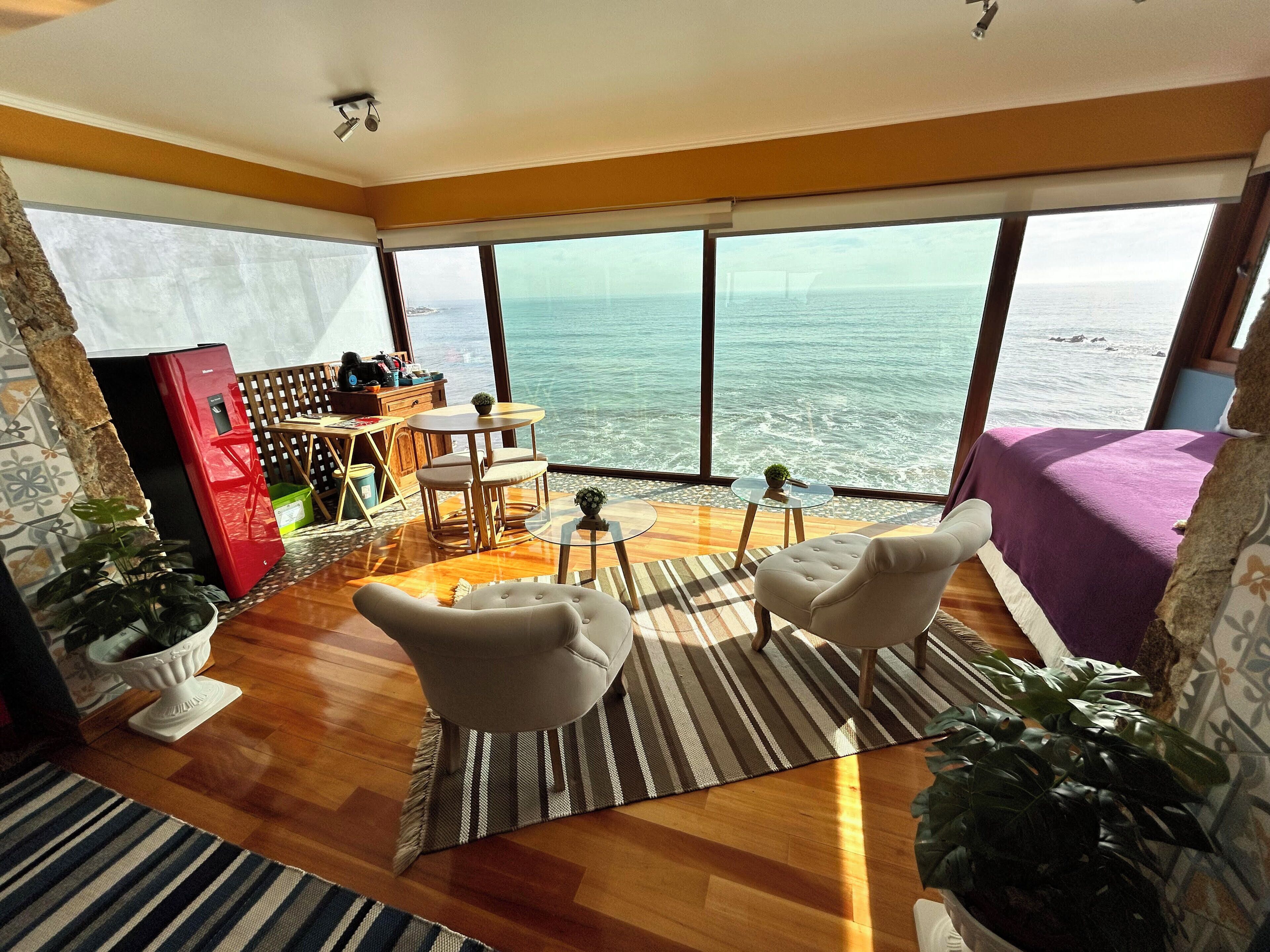 Una habitación amplia con piso de madera, un comedor con mesa y sillas, un sofá y un gran ventanal con vista al mar.