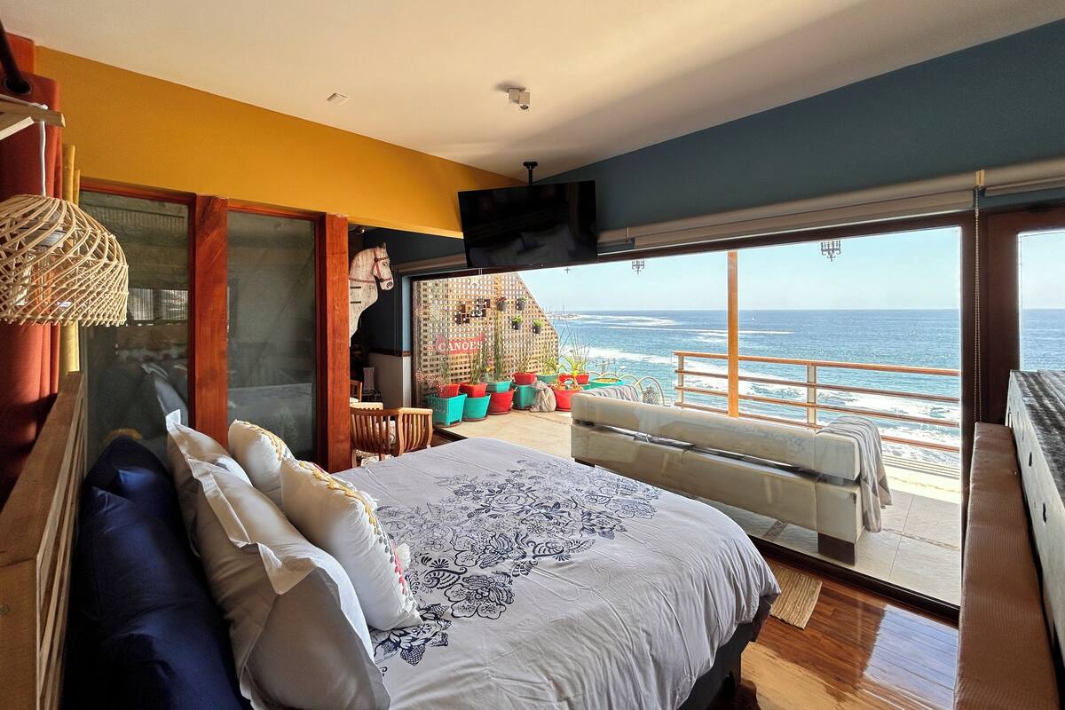 A bedroom with a large bed, a view of the ocean, and a balcony with potted plants.