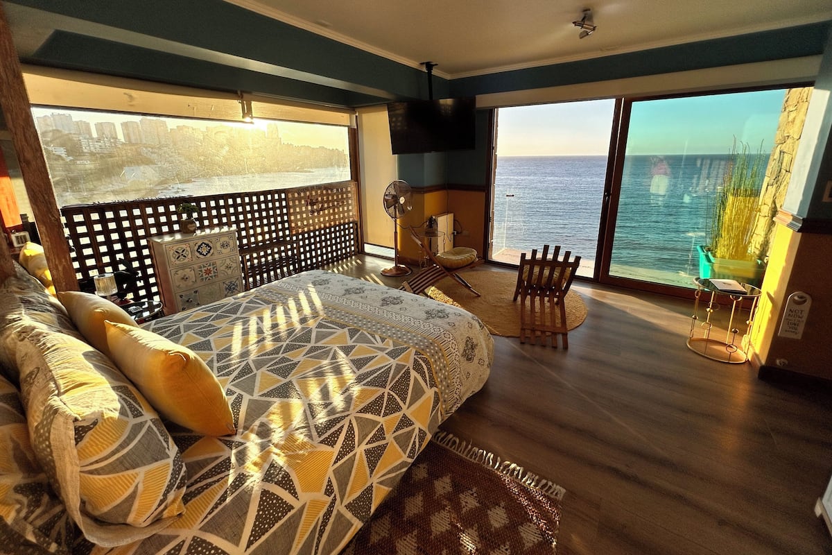 A bedroom with a large bed, a chair, a small table, and a view of the ocean through large windows.