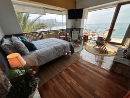 Een slaapkamer met een groot bed, een televisie, een balkon met uitzicht op zee, en een zithoek met stoelen en een tafel.