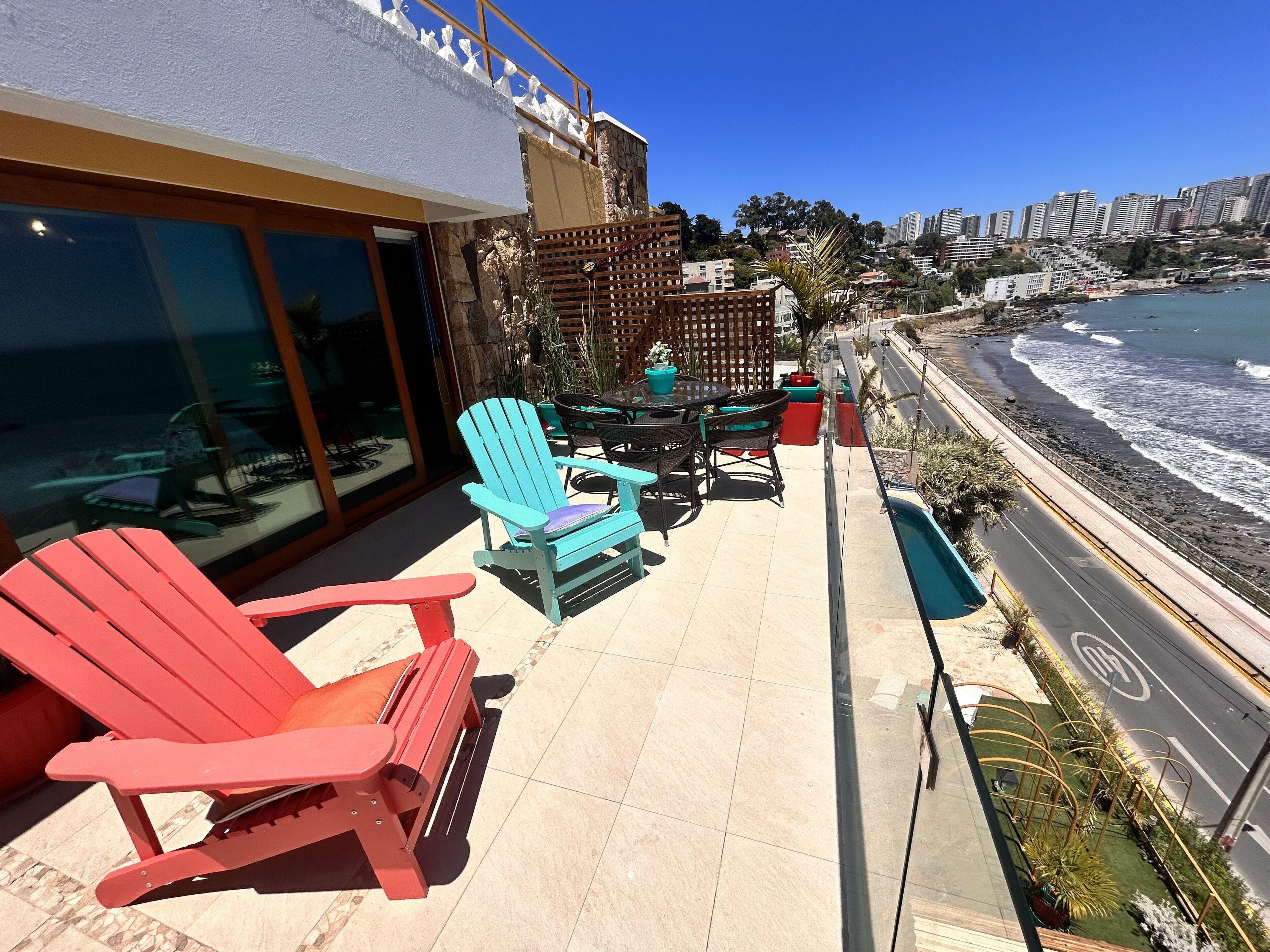 A balcony with red and blue Adirondack chairs, a table, and potted plants. A road runs parallel to the balcony, and a beach with waves is visible in the background.