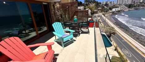A balcony with red and blue Adirondack chairs, a table, and potted plants. A road runs parallel to the balcony, and a beach with waves is visible in the background.