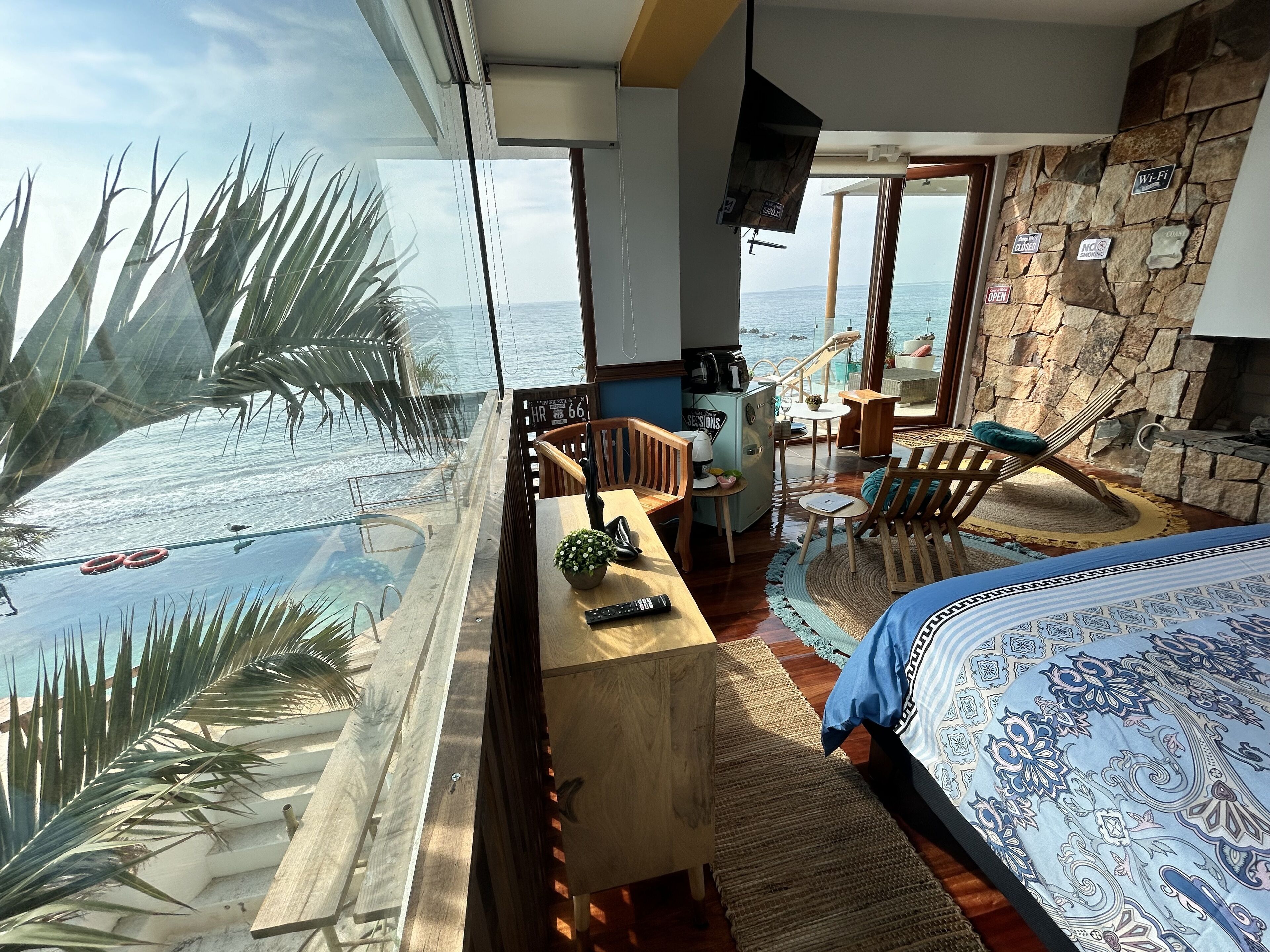 Una habitación con un ventanal grande con vistas al mar, una cama con ropa de cama estampada, un escritorio de madera, una silla y una mesita con una planta.