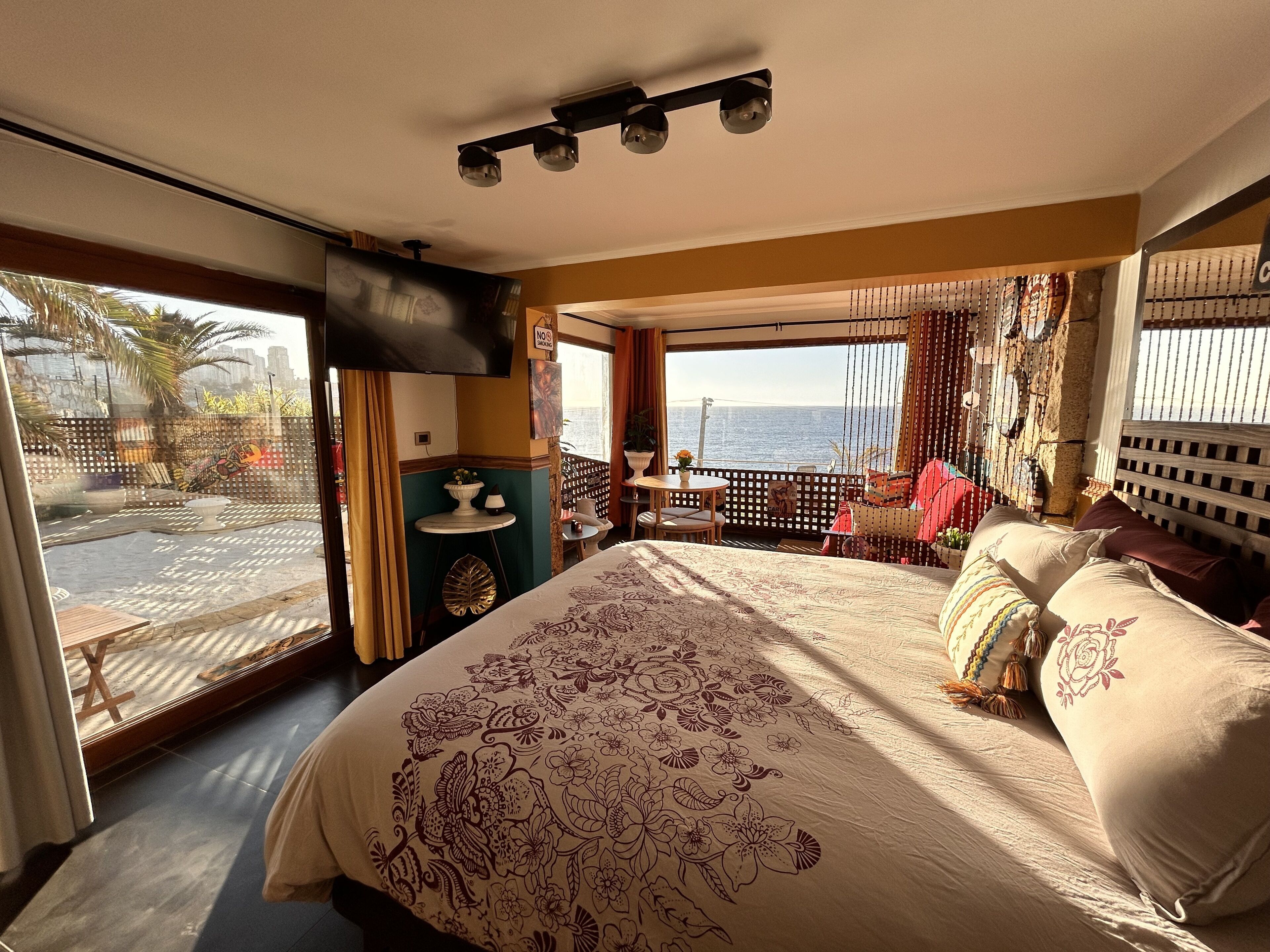 A bedroom with a large bed, a TV mounted on the wall, a balcony with a view of the sea, and a small table with chairs.