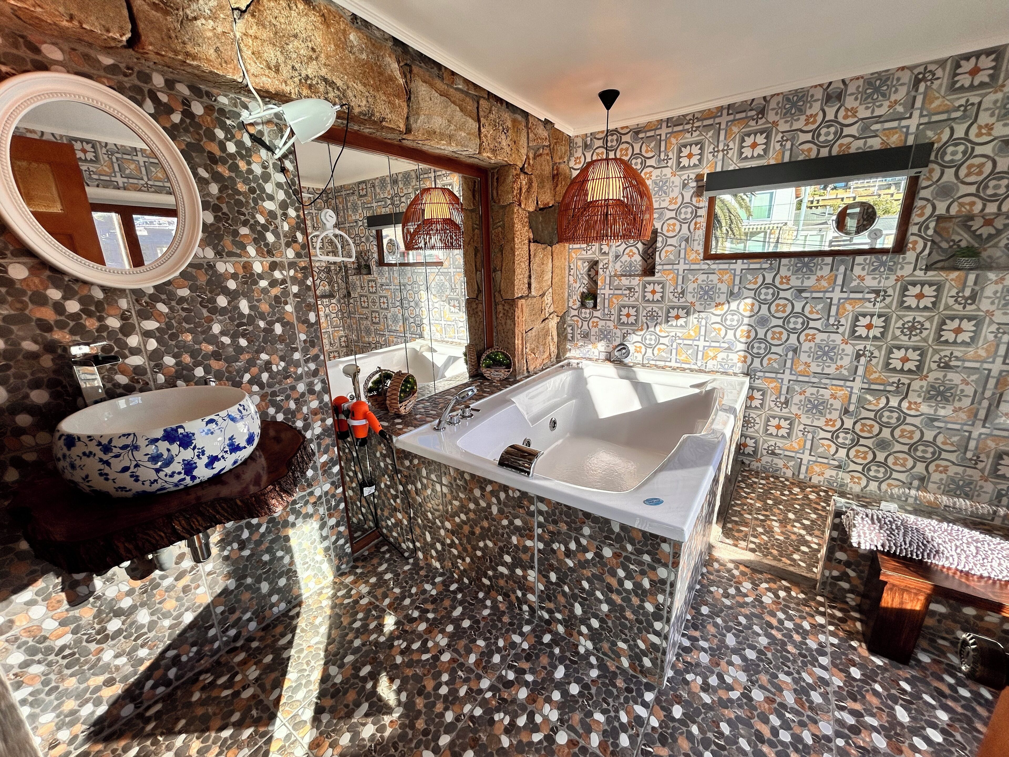 A bathroom with a large oval sink, a freestanding bathtub, and a mirror. The walls are decorated with patterned tiles, and there is a window providing natural light.