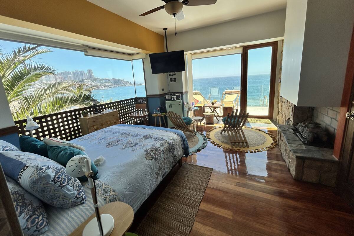 A bedroom with a bed, a desk, a chair, a TV, and a balcony with a view of the ocean.