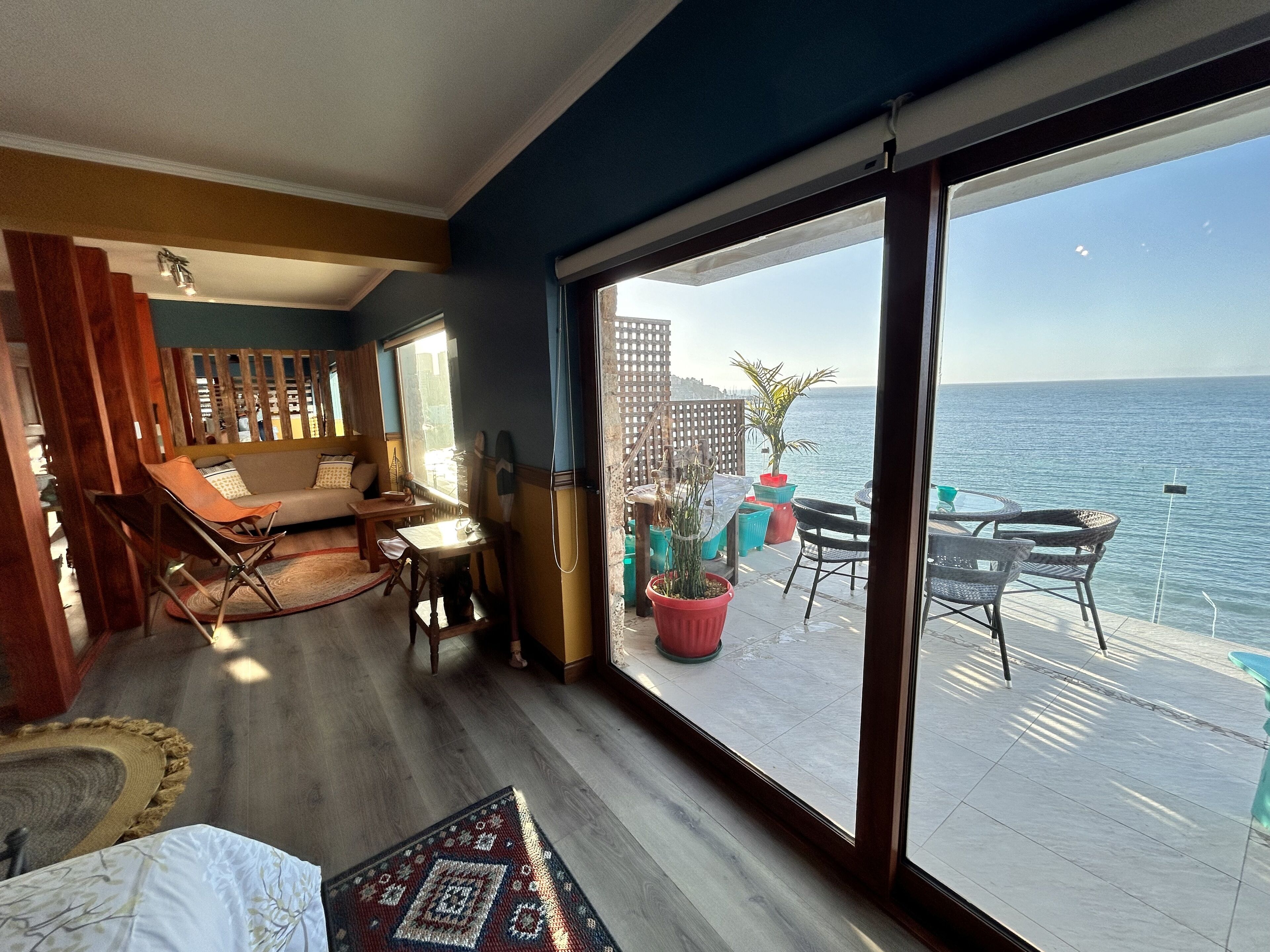A room with a large sliding glass door leading to a balcony with a view of the sea. Inside, there is a sofa, a dining table, and a small seating area with chairs.