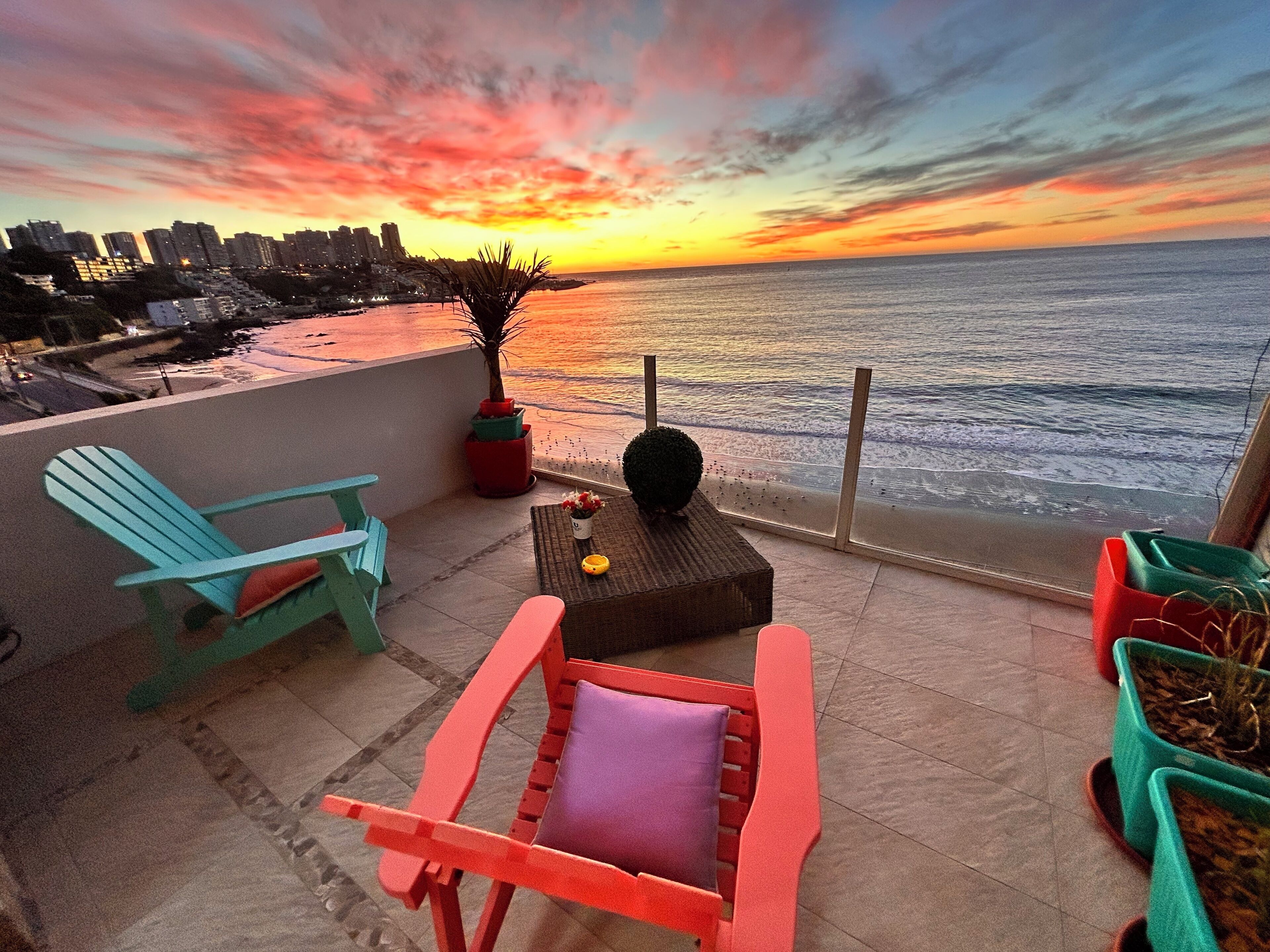 A terrace with a view of the ocean at sunset, featuring a chair, a small table, and potted plants.