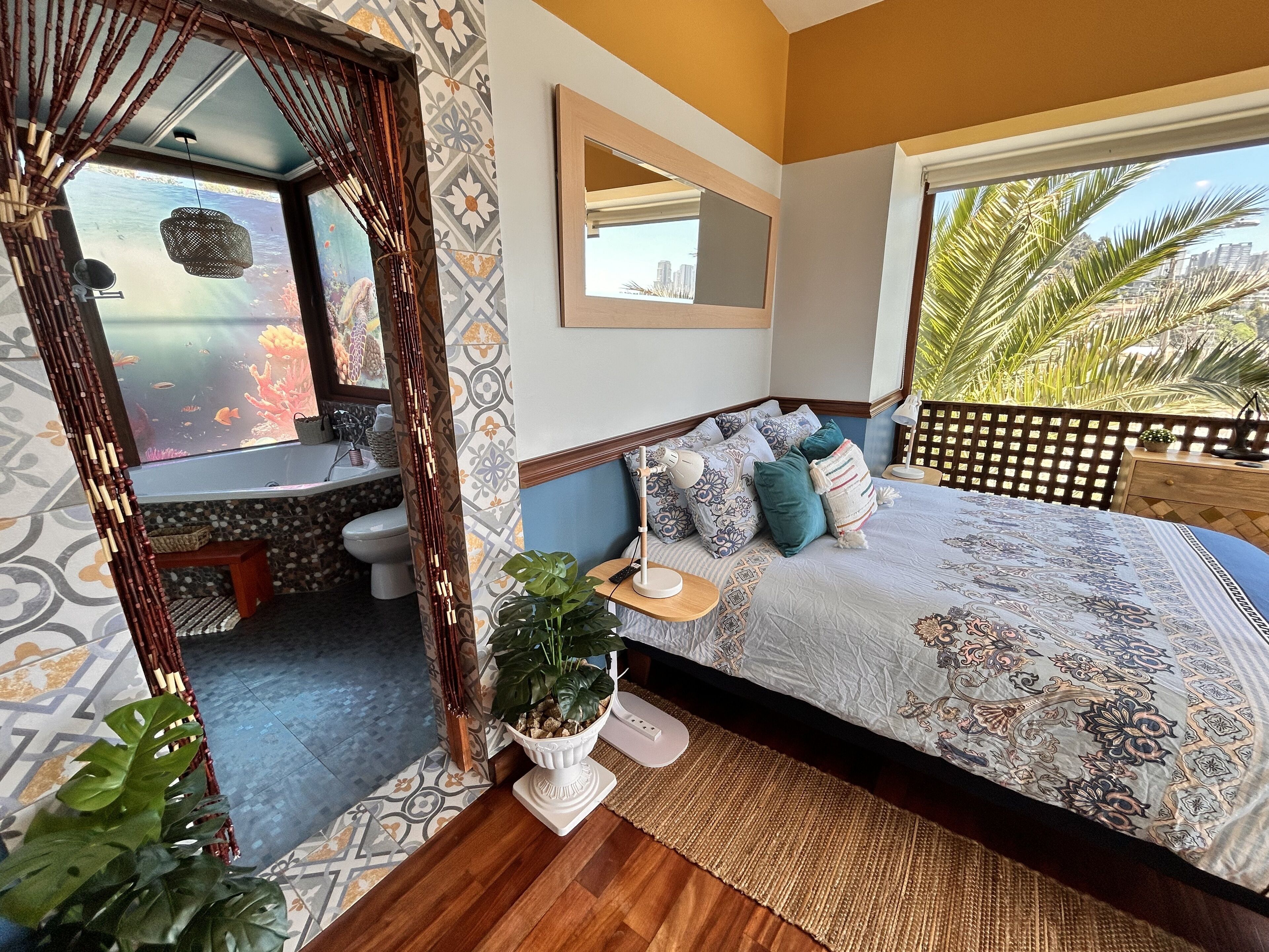 A bedroom with a large bed, a view of the city, a bathroom with a bathtub, and a balcony with plants.