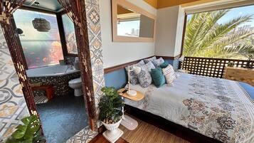 A bedroom with a large bed, a view of the city, a bathroom with a bathtub, and a balcony with plants.