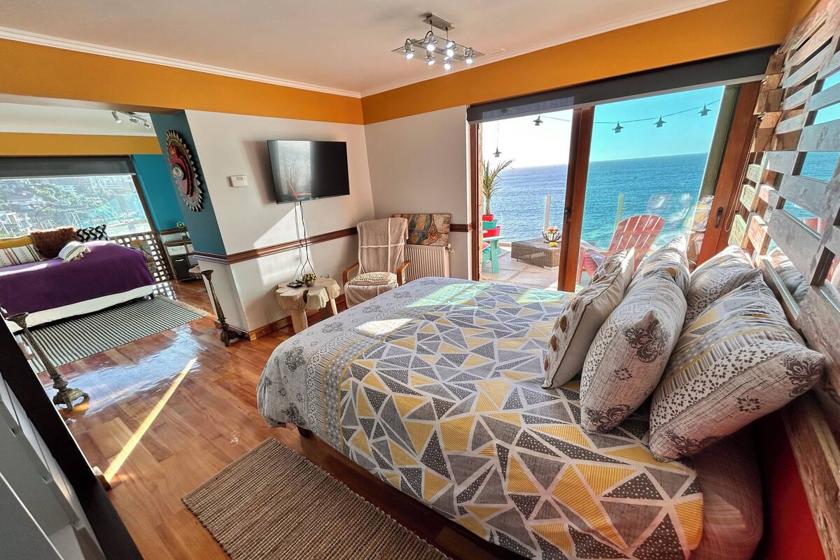 A bedroom with a large bed, a TV, and a balcony with a view of the sea.