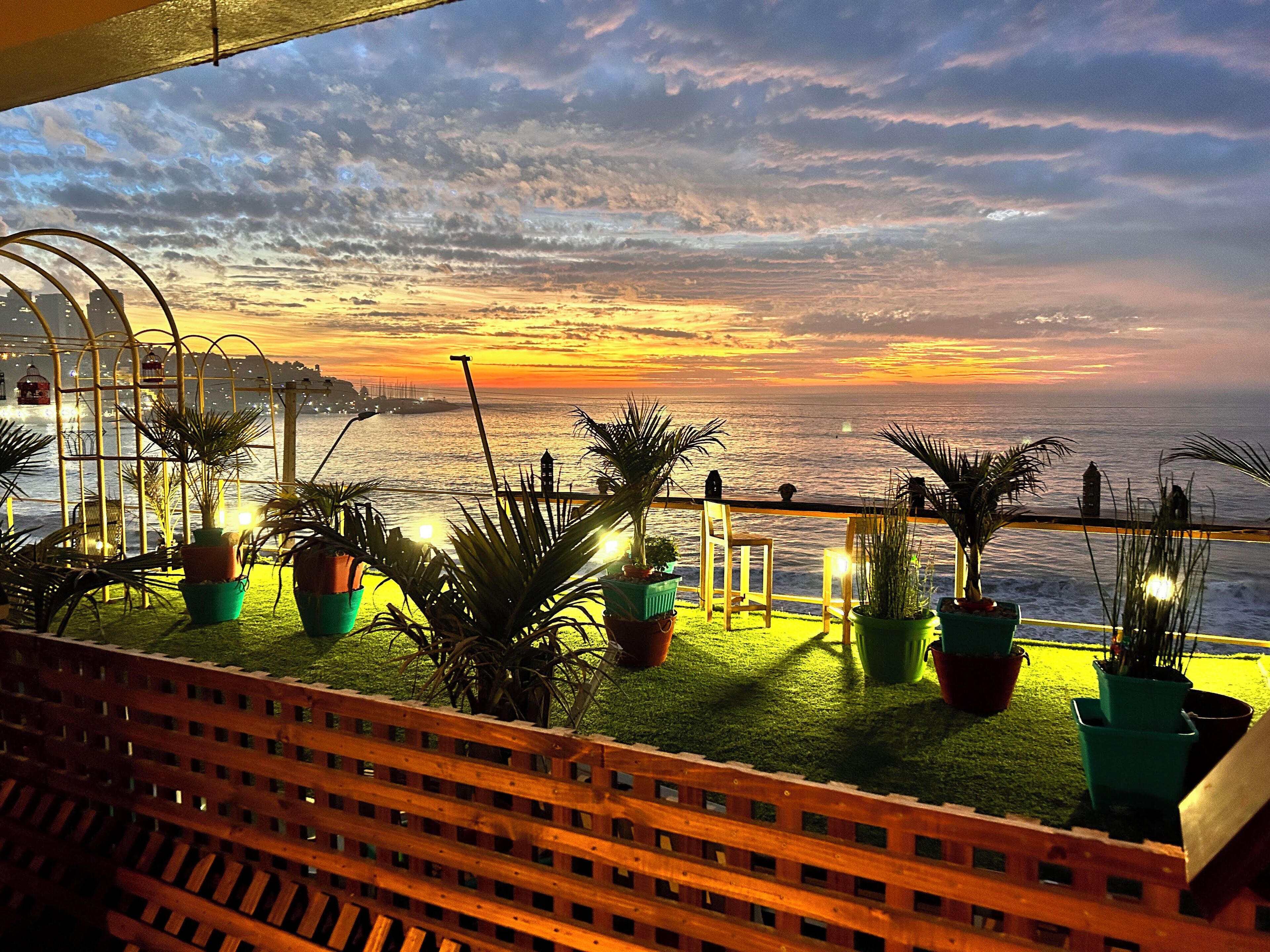 A balcony with potted plants, a wooden railing, and a view of the sunset over the ocean.