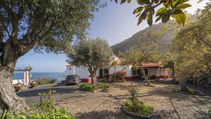 Ein Pflasterhof mit einem weißen Gebäude, Olivenbäumen und Blick auf das Meer.