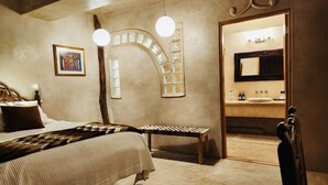 A bedroom with a bed, a bench, a mirror, and a door leading to a bathroom. - Alegria Suites Centro Historico  (Todos Santos)