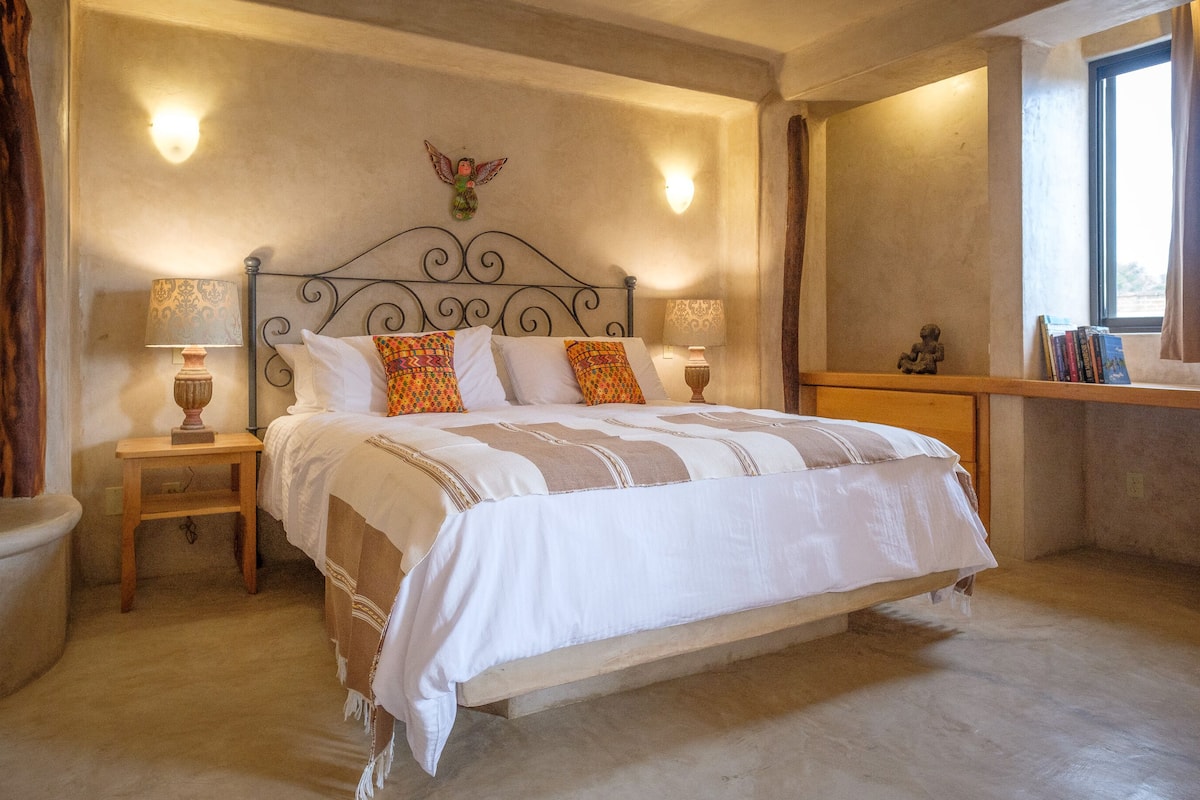 Een slaapkamer met een groot bed, twee nachtkastjes met lampen, een houten bankje, een wandplank met boeken en een raam met gordijnen.