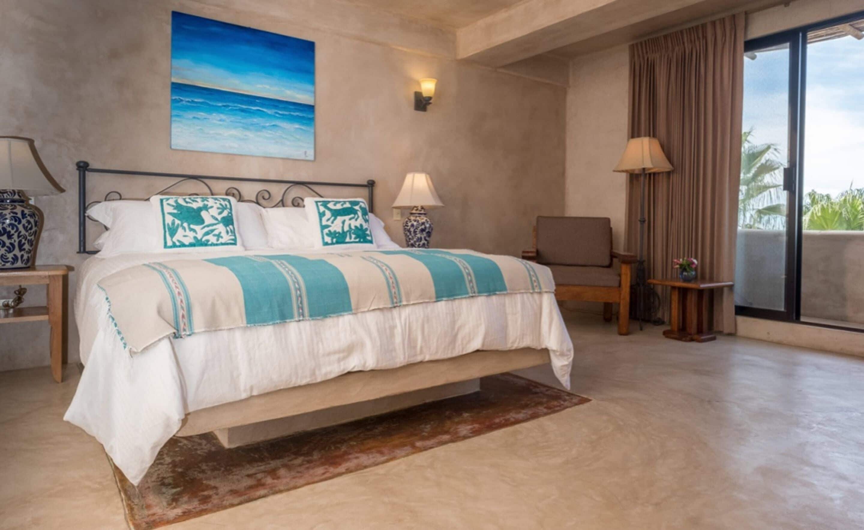 Ein Schlafzimmer mit einem Bett, einem Nachttisch, einem Sessel, einer Lampe und einem Strandbild.