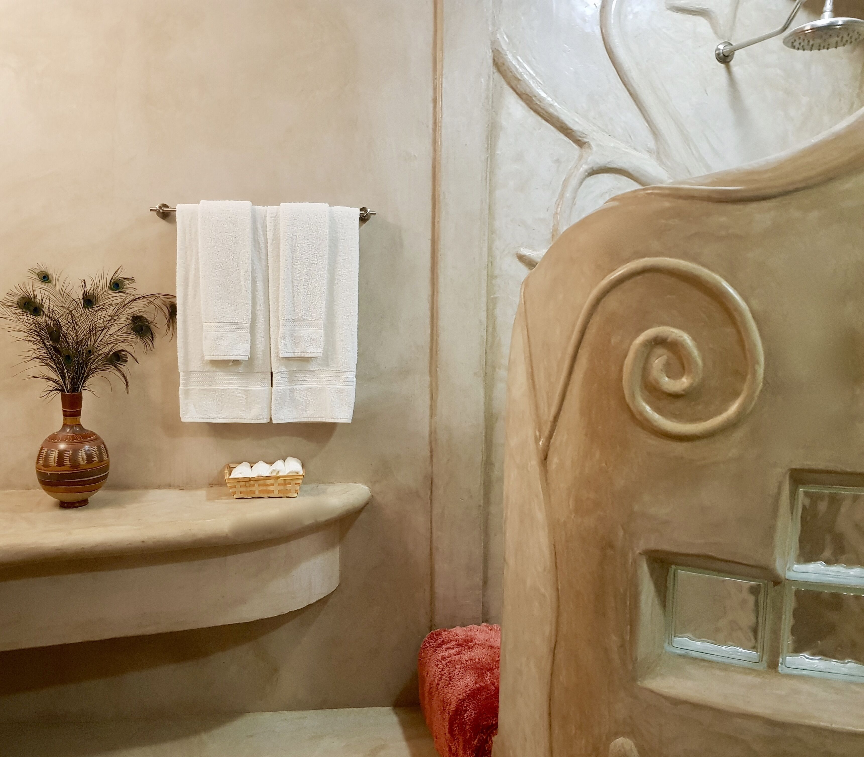 A bathroom with a beige wall, a towel rack with two towels, a decorative vase with dried flowers, and a small basket of toiletries.