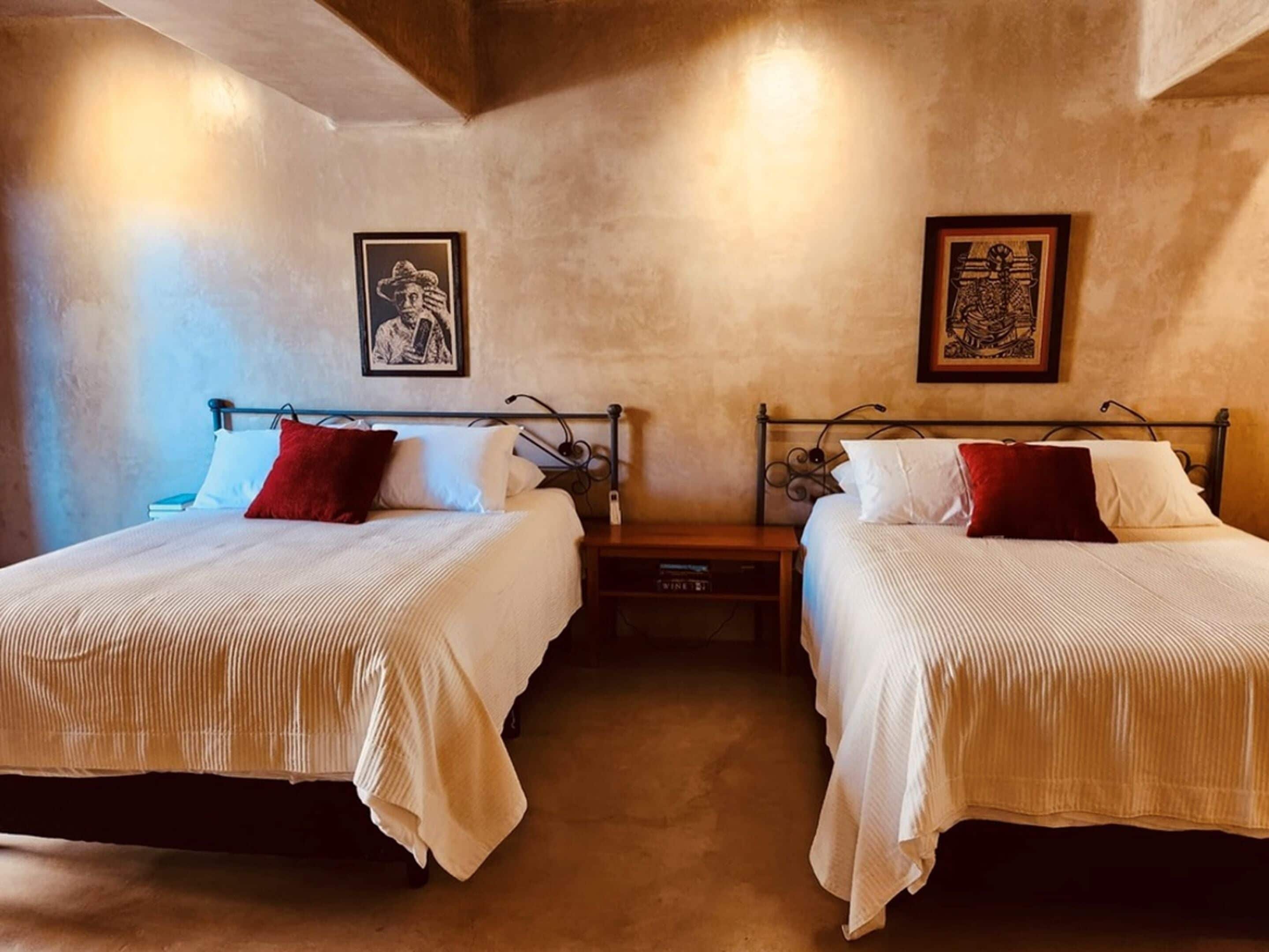 Une chambre d’hôtel avec deux lits, chacun agrémenté de draps blancs et d’oreillers rouges. Les murs sont ornés de tableaux encadrés, et on y trouve une table de chevet en bois ainsi qu’une petite table.