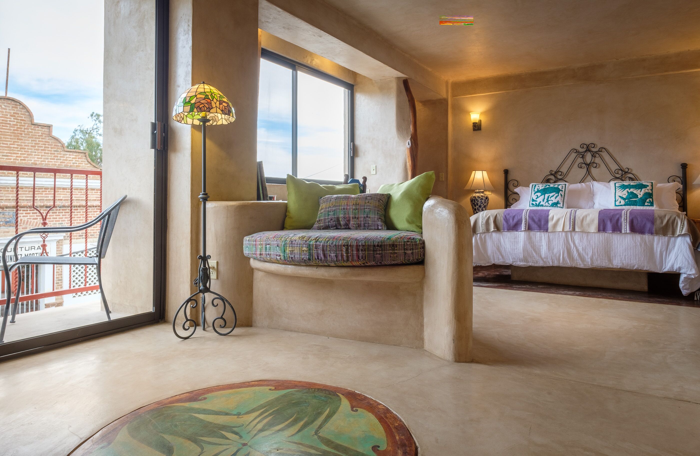 A bedroom with a bed, a bench with cushions, a floor lamp, and a view of the outside.