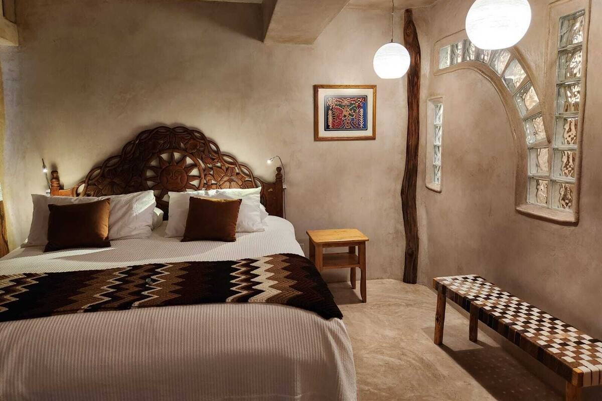 Een slaapkamer met een groot bed, een houten hoofdeinde, een bankje, een kleine tafel, een wandplank en een decoratief raam.