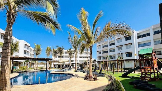 A resort pool area with palm trees, a children's play structure, and a building with multiple balconies. - Almarena Apartments Coto Coral (Mazatlán)