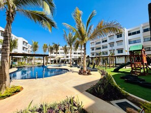 Zona de piscina en un resort con palmeras, un área de juegos infantiles y un edificio con varios balcones.