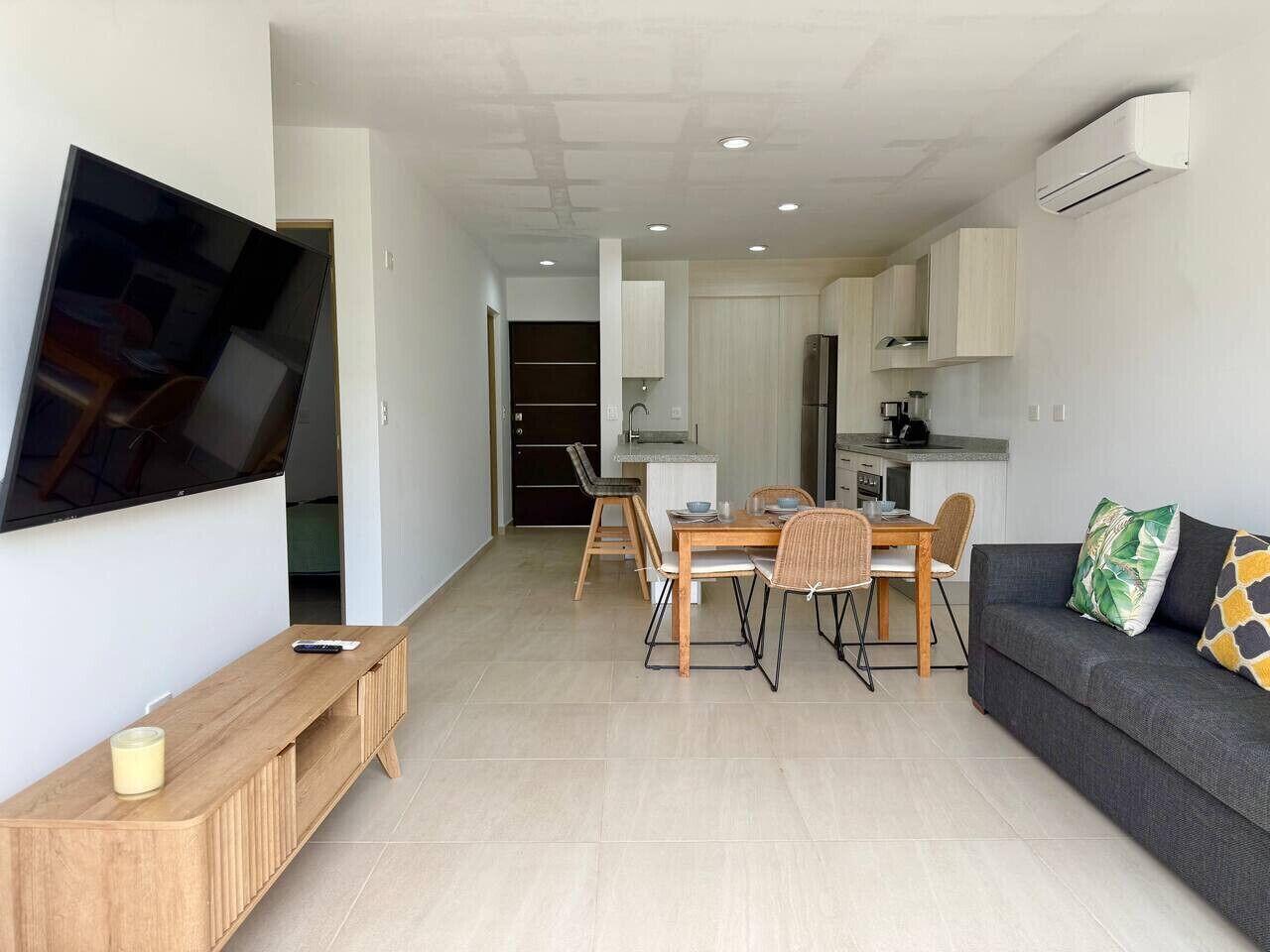 Ein modernes Wohnzimmer mit Flachbildfernseher, einem Holzkonsolentisch, einem Essbereich mit Tisch und Stühlen sowie einem grauen Sofa mit dekorativen Kissen.