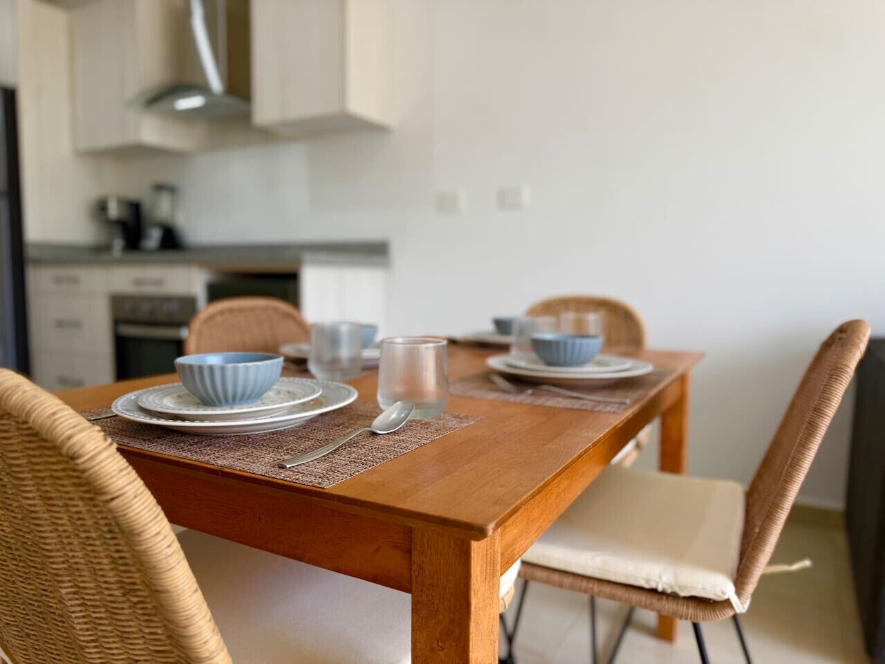 A wooden dining table with blue bowls, a glass, and a spoon. There are wicker chairs and a cushioned seat. A kitchen is visible in the background.
