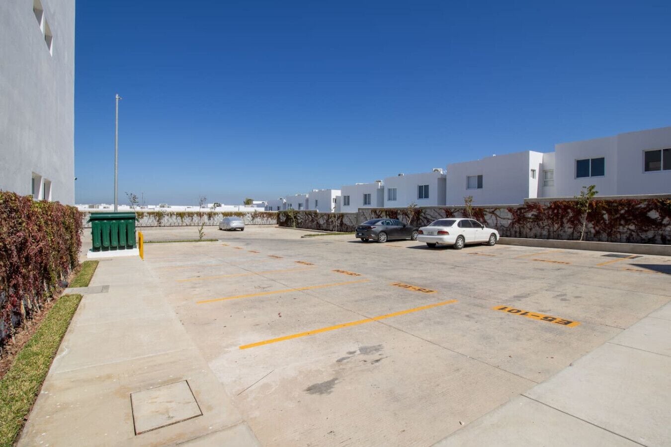 Ein Parkplatz mit markierten Stellplätzen, ein grüner Müllbehälter und eine Reihe moderner Gebäude mit Fenstern.