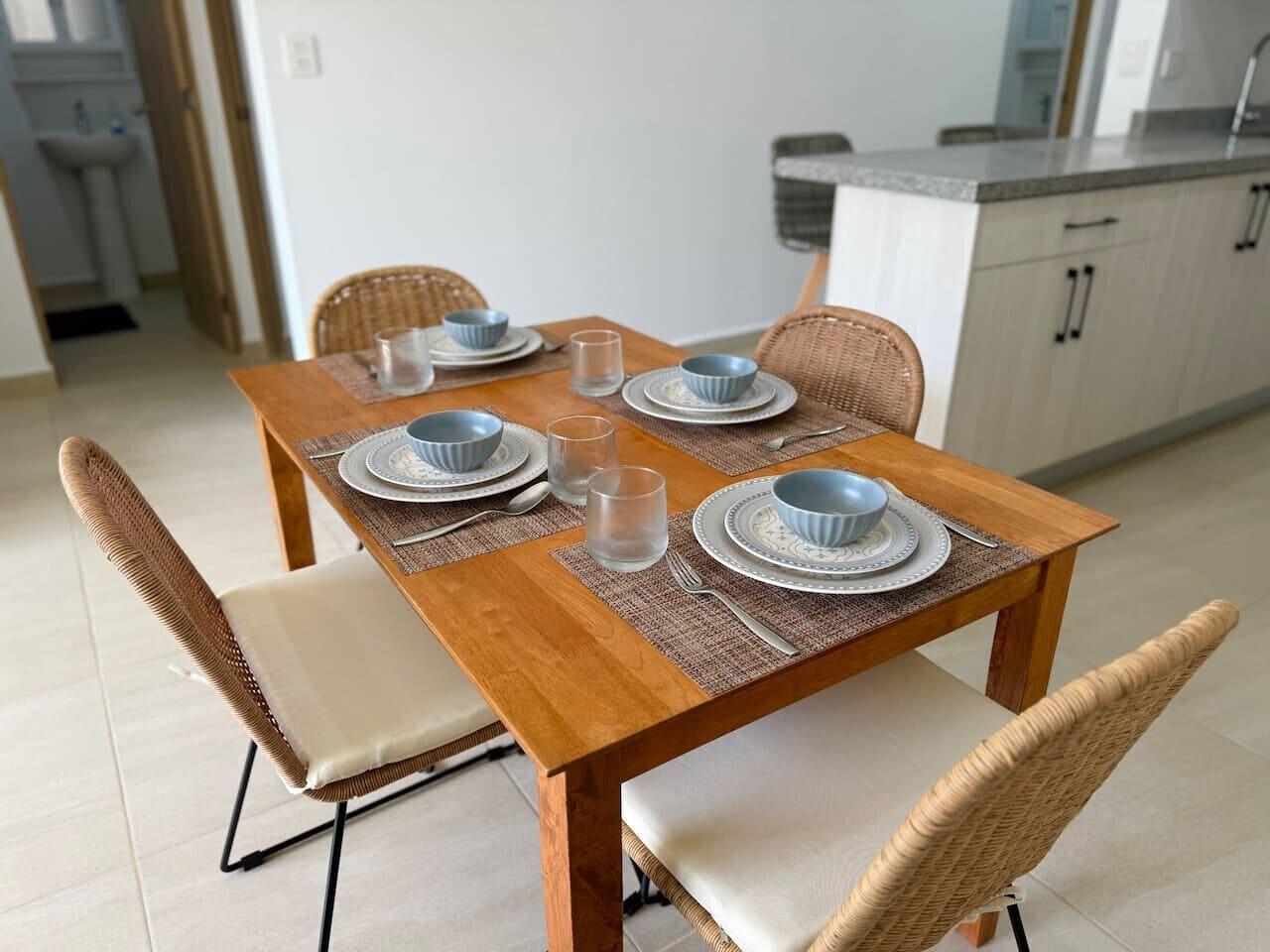Une table à manger dressée avec des assiettes, des bols, des verres et des couverts, entourée de chaises en osier.