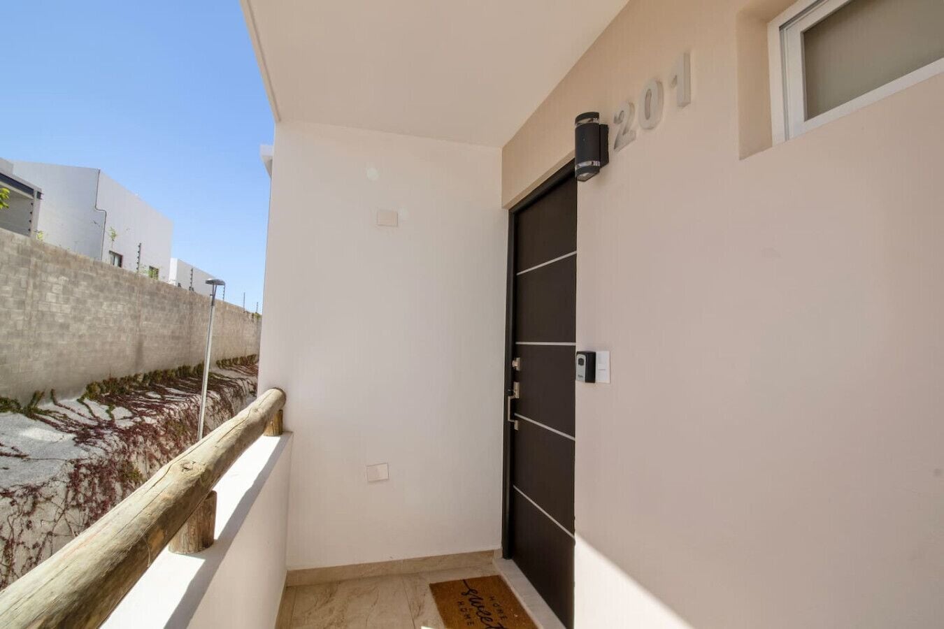 Un balcon étroit avec une balustrade en bois, une porte noire dotée d’un clavier pour l’ouverture, et une petite fenêtre sur laquelle est inscrit le numéro « 201 ».