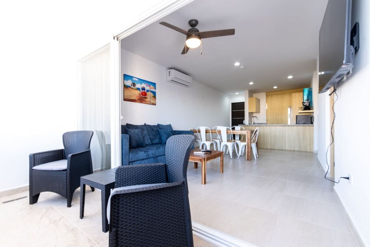 A modern living area with wicker furniture, a ceiling fan, and a kitchen in the background.