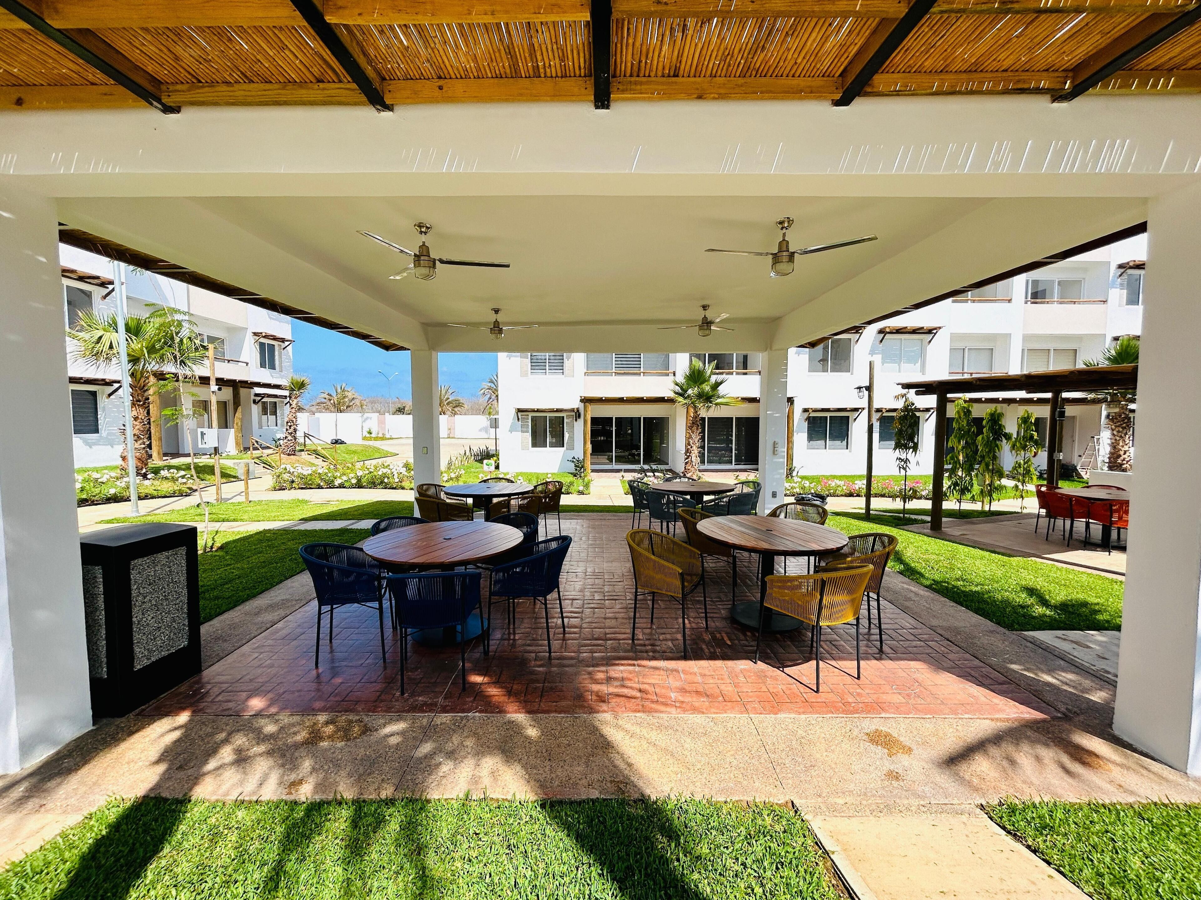 Un patio techado al aire libre con varias mesas y sillas, un ventilador de techo y vistas de edificios circundantes.